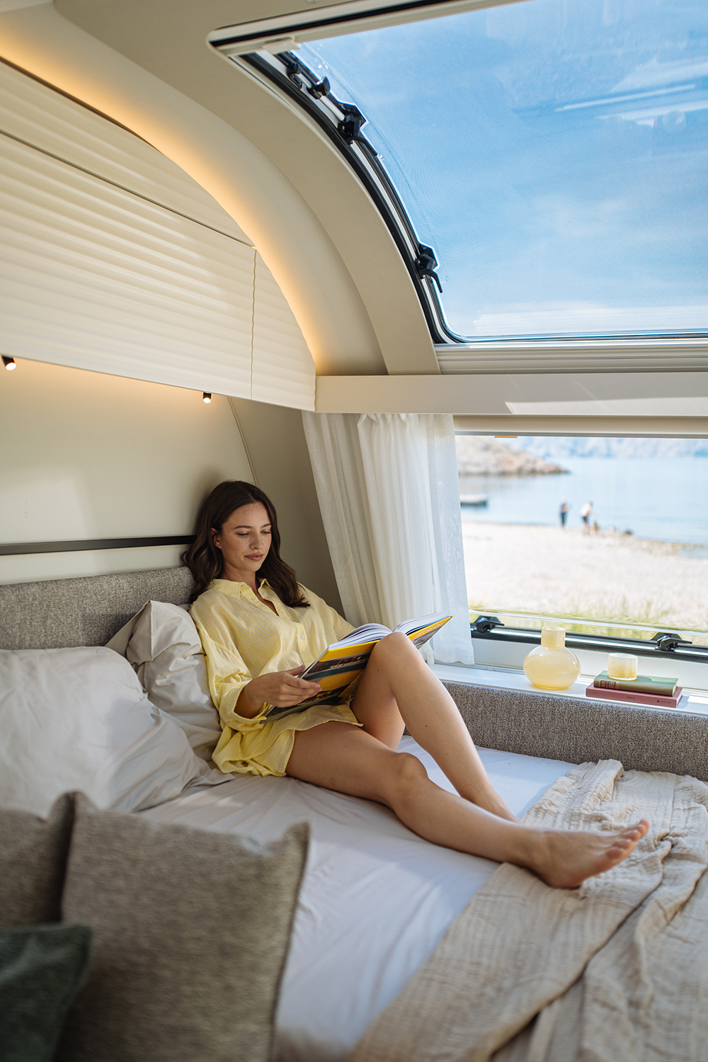 Eine Frau sitzt auf dem Elternbett unter dem Panoramafenster im ADRIA ADORA Wohnwagen und liest ein Buch.