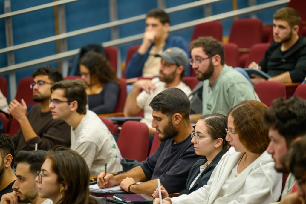 A diverse group of students sitting in an auditorium or lecture hall taking notes during a class or presentation.