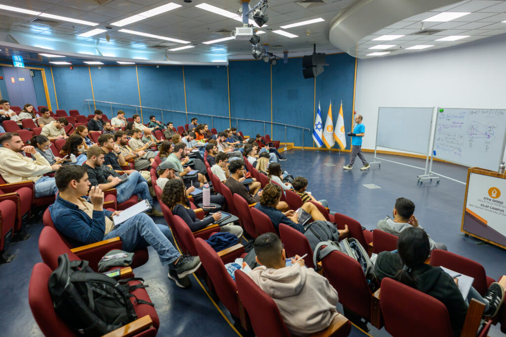 Large lecture hall filled with students seated in red chairs listening to a presenter near a whiteboard