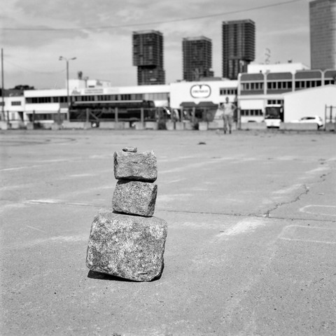 The black and white image shows a small stack of four uneven stones balanced on a flat, empty concrete surface. In the background, there are industrial buildings and several tall, modern towers, giving a contrast between the simple stones and the urban environment.