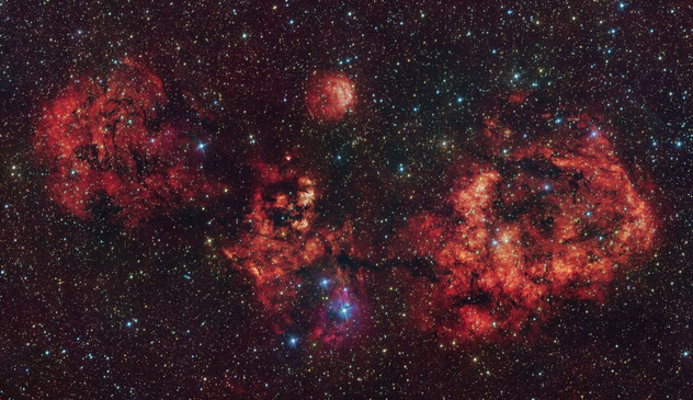 A telescopic view of a cosmic cloud against a dark starry sky. The cloud resembles a flying bat, spanning its wings. The cloud is mostly red, with some areas partially blocked by dark dust clumps.