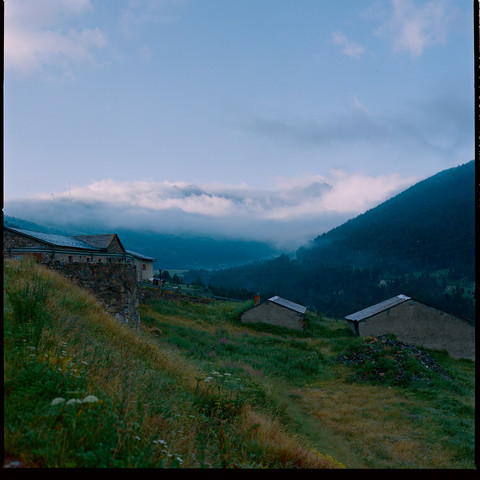 Traditional stone buildings nestle in the lush valley, offering a glimpse of the distant, hazy mountain range beneath a cloudy sky.