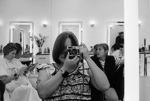 a scan of a black and white film photo of me taking a self portrait using a film camera. the setting is at a hair salon in the upper east side in new york, with two ladies in the background sitting in front of mirrors. i am holding up a camera and pointing it into the mirror