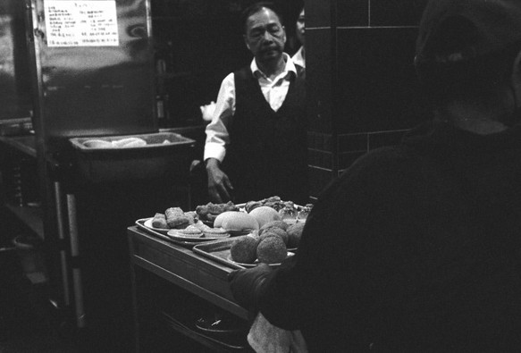a scan of a black and white film photograph showing a dimsum cart with some buns. in the background, a waiter looks on. there are chinese menus or notices in the background.