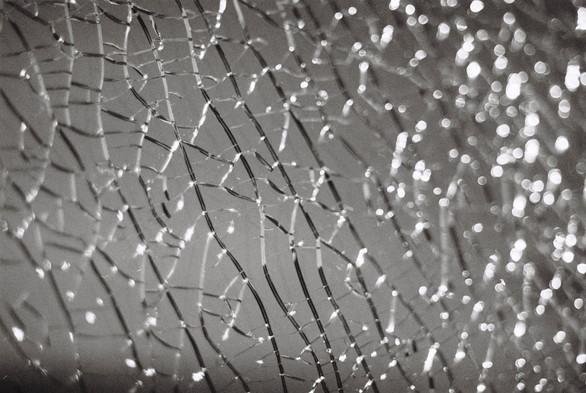 Close-up black-and-white photograph of a shattered glass surface. A web of cracks covers the entire surface like glimmering silver threads. The reflections in the background almost look like the sun shining on waves in the sea. Pretty, but broken.