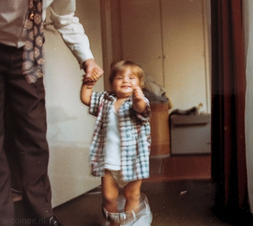 A vintage photograph featuring a toddler holding hands with his father. The toddler is very happy, smiling broadly and looking directly at the camera. 

He is wearing an open plaid shirt with some red, blue and white tones over a white undershirt. 

He is also wearing a pair of oversized, light-colored trousers which are about to fall on the ground.