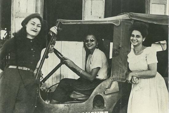 Patria, Minerva and Maria Teresa Mirabal. One is at the wheel of a jeep and the other two stand either side of her.
