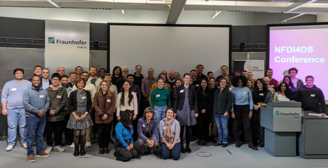 Attendees of the 2025 NFDI4DataScience conference lined up in front of the stage of the large conference room at Fraunhofer Fokus in Berlin.