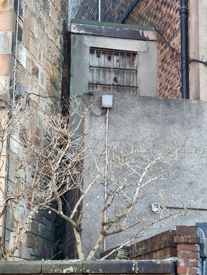 A small concrete box with a barred and broken window wedged between two buuldings.