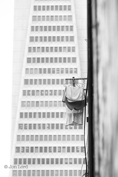 This is an abstract black and white street photo in portrait format of some washing drying from a window with an iconic high-rise in the background. San Francisco (2015).

In the foreground and stretching away from the camera along the right margin of the image is the pale wall of a building, heavily blurred out of focus. Attached to the wall are some loose cables and what might be a black drain pipe running vertically up the wall of the building. To the right of centre, and in focus, is a short metal pole of about a metre long protruding from the wall at right angles, with a small amount of laundry hanging from metal coat-hangers and out to dry. In the background and slightly blurred out of focus is the facade of a tall, triangular shaped, white office block. Only part of the middle levels are in view, the lower floor having about seventeen windows stretching across, with the building tapering to nine windows across on the upper floor. There are many floors out of view below and a few more floors out of view above the image. 

The tapered building is the Transamerica Pyramid (also know as the Transamerica Tower and Transamerica Building) in San Francisco's finanical district. The drying laundry: I cannot comment on.
