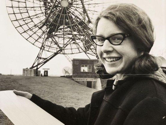 Jocelyn Bell Burnell in the 1960s, in front of a radio telescope. she is a white woman with dark hair, wearing a sensible duffle coat given how cold it must have been out there.
