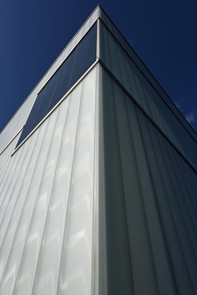 Color photo looking up at the edge of a structure clad in vertically oriented, translucent glass planks. There is a horizontally oriented window set into the upper reaches of the wall on the left. The sky above the structure is a deep blue.