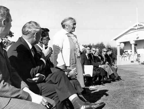 Iriaka Rātana standing at an outdoor meeting, surrounded by men sitting in suits. She is a Māori woman with white and grey hair.