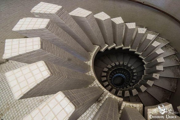 A spiral staircase with white tiled steps and vertical tiled columns, viewed from above, creating a geometric, circular pattern.
