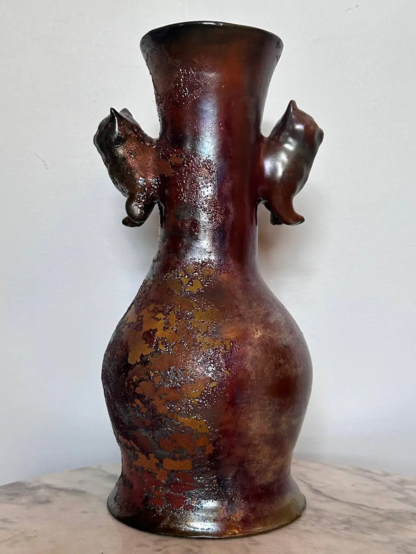 A red, brown and copper raku vase with cats on the neck of the vase, on a white background
