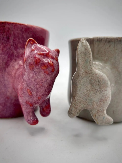 A red cat mug with cat shaped front of body handle. A cream coloured mug with a cat shaped handle of the cat's back end.