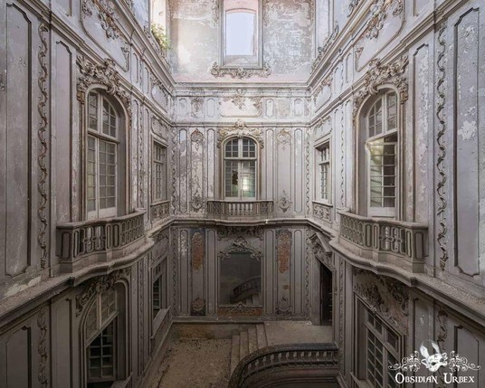 Abandoned grand interior with ornate moldings, peeling paint, tall windows, and a curved staircase, showing signs of decay and neglect.