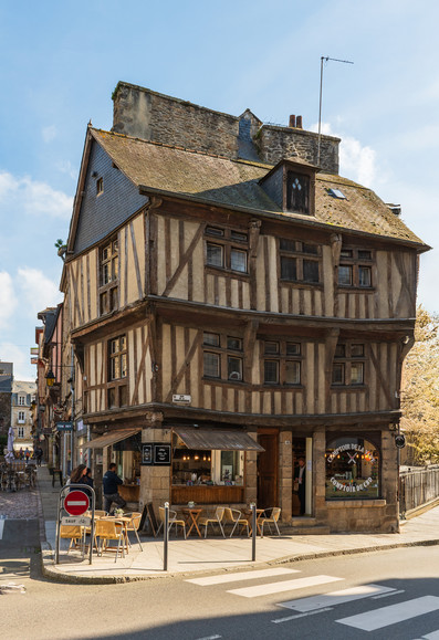 Das Bild zeigt ein malerisches, mehrstöckiges Fachwerkhaus an einer Straßenecke. Vor dem Geschäft stehen Tische und Stühle, die zum Verweilen einladen. Auf der Straße ist ein Zebrastreifen zu sehen. Die Architektur des Hauses ist traditionell und charmant. Im Hintergrund erstreckt sich eine Gasse mit weiteren Gebäuden und ein paar Passanten. Ein Verkehrsschild, das Fahrräder verbietet, steht am Straßenrand. Der Himmel ist blau und der Tag sonnig.