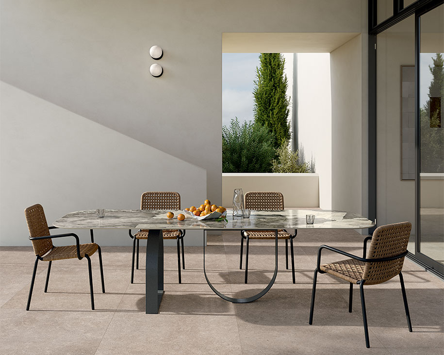 Gartentisch mit grau geäderter Steinzeugplatte in Marmoroptik und Metallgestell, geflochtenen Stühlen und einem modernen architektonischen Hintergrund.