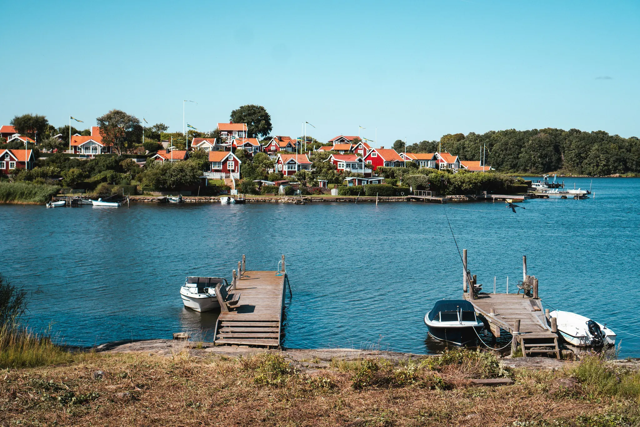 A scenic view of a coastal village featuring numerous red-roofed houses amidst green trees, bordering a calm blue sea.…