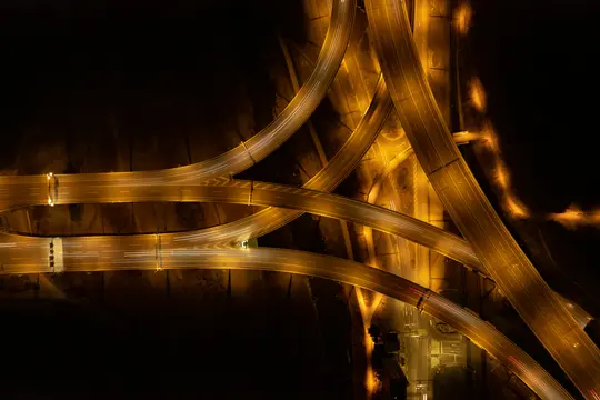 An aerial, top-down view of a complex highway interchange at night, with multiple roads curving and crossing. The roads are…