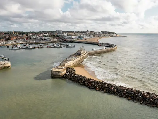 En flygfoto över en hamn med många förtöjda båtar, en lång, böjd stenpir, en sandstrand och en stad längs kusten.