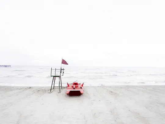 A red lifeguard stand with a dark red flag and a red paddle boat sit on a light sandy beach next to a grey ocean.