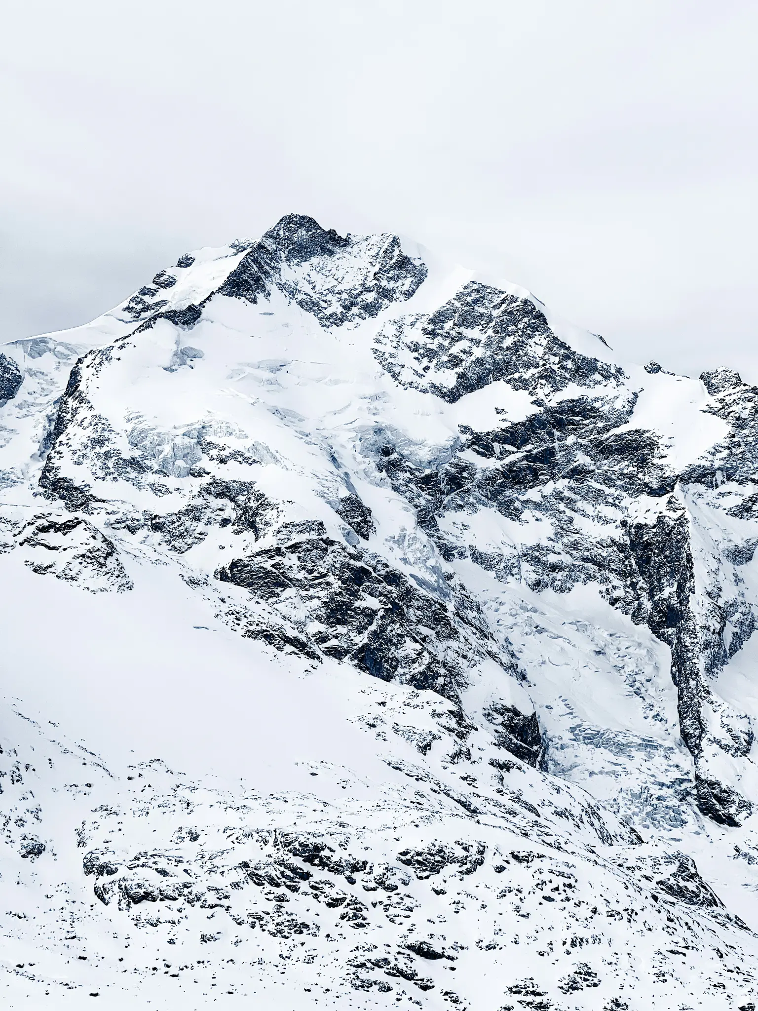 En tornande bergstopp täckt av tjock vit snö och mörkgrå sten, med synliga glaciärer och en molnig himmel.