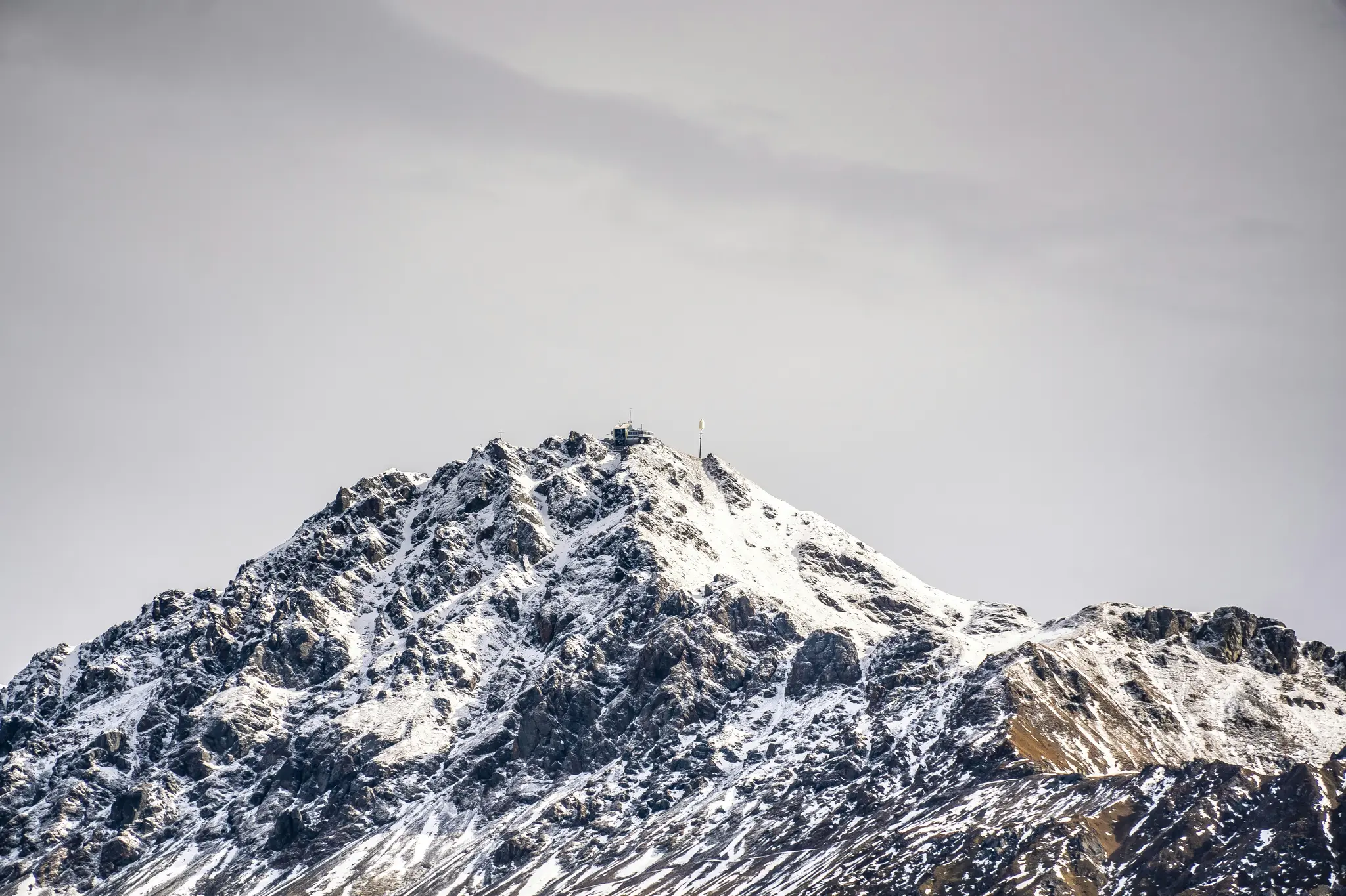 En karg, snötäckt bergstopp reser sig under en grå, molnig himmel. En liten byggnad och en antenn syns.