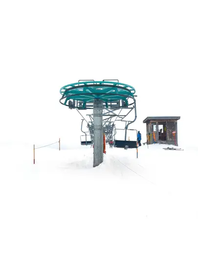 En turkis og grå skiliftmekanisme står fremtrædende på en stor hvid sneflade med en lille træhytte til højre.
