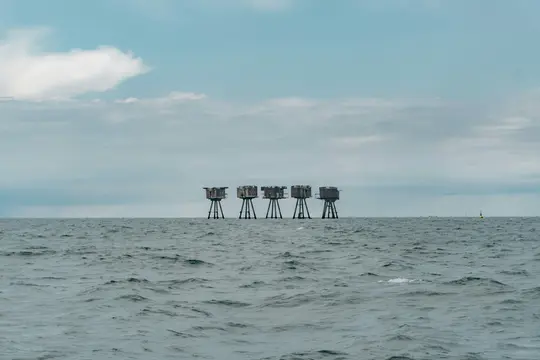 Ett brett landskapsfoto visar fem väderbitna havsfästningar, var och en på fyra pålar, uppradade vid horisonten av ett grått