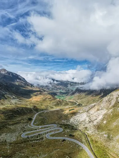 En flygvy visar en slingrande väg som skär genom en vidsträckt bergdal under en molnig himmel. Gröna och steniga sluttningar…