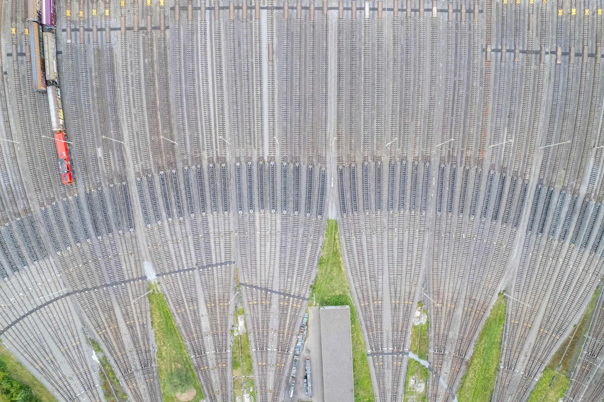 Una toma aérea muestra un vasto patio de maniobras de trenes con innumerables vías paralelas. Un tren rojo es visible en una