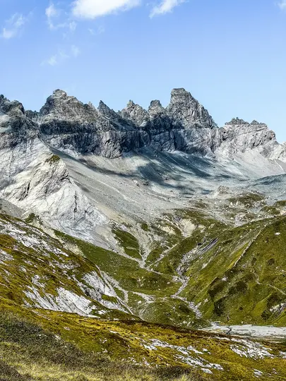 A wide landscape view of a mountain range with sharp, rocky gray peaks under a light blue sky with white clouds. The lower…