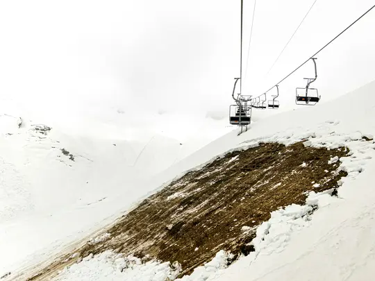 En tom skidlift sträcker sig över ett vidsträckt, snötäckt berg. En stor fläck av brun jord är synlig på sluttningen.
