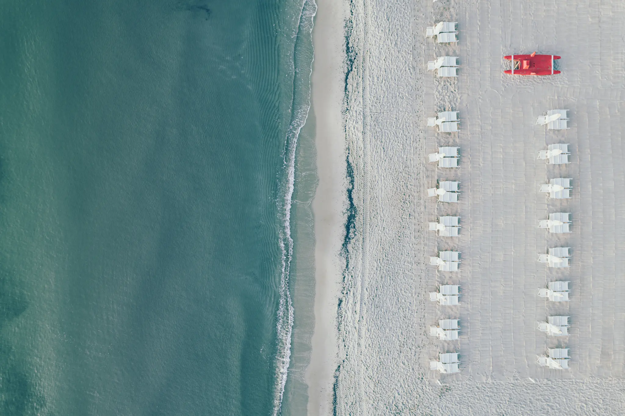 An aerial, top-down view of a tranquil beach. Turquoise water meets a white sandy shore with gentle waves. Two parallel…