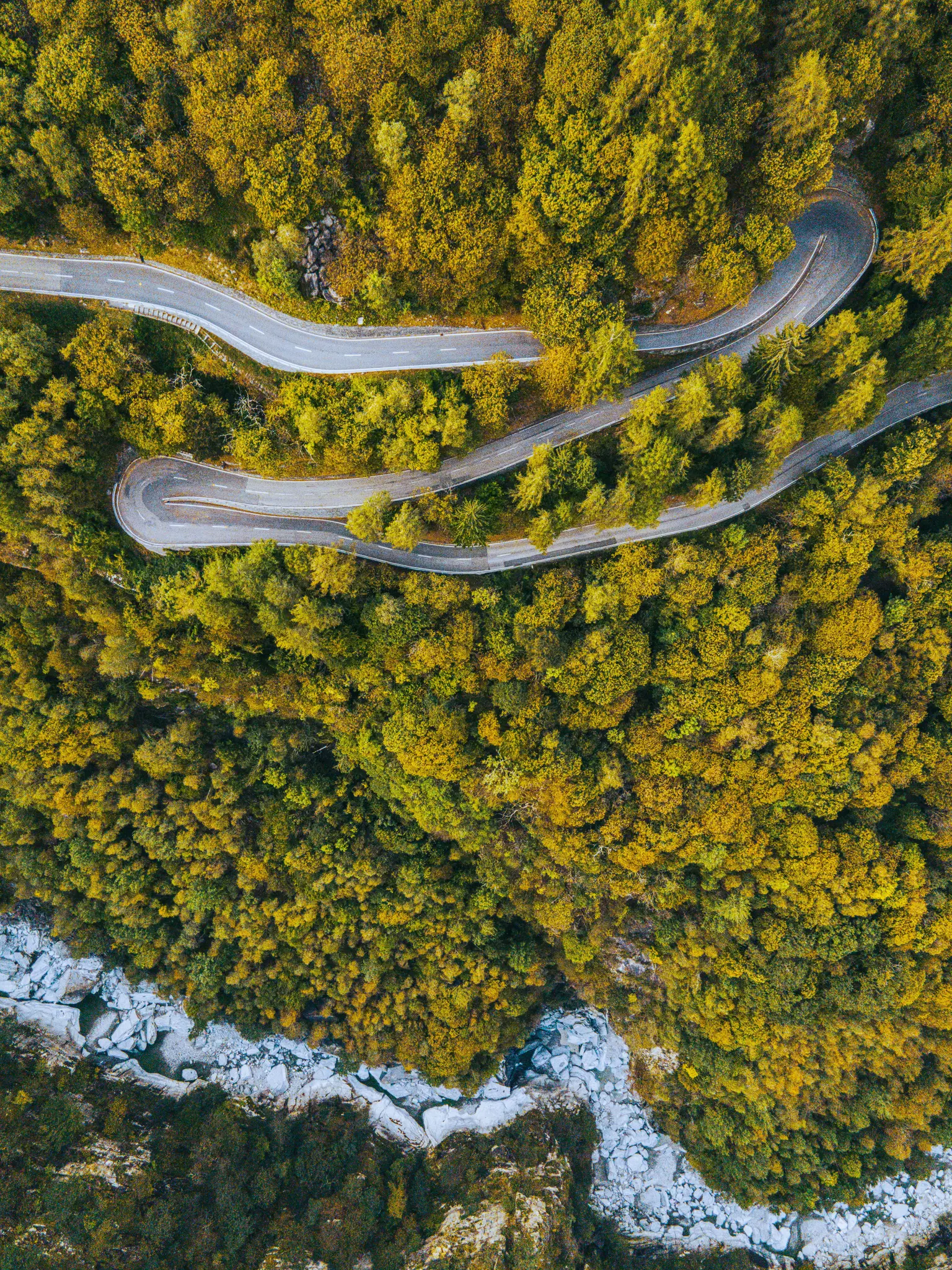 An aerial view shows a grey winding road snaking through a dense forest of green and yellow autumn trees. A rocky riverbed…