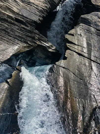 En närbild, vertikal bild av ett vattenfall som forsar nerför en mörk, lagrad stenig ravin, med solljus som reflekteras från