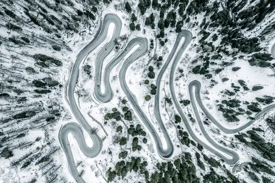 En flygvy visar en grå, slingrande väg som slingrar sig genom ett snötäckt berglandskap med mörkgröna barrträd.