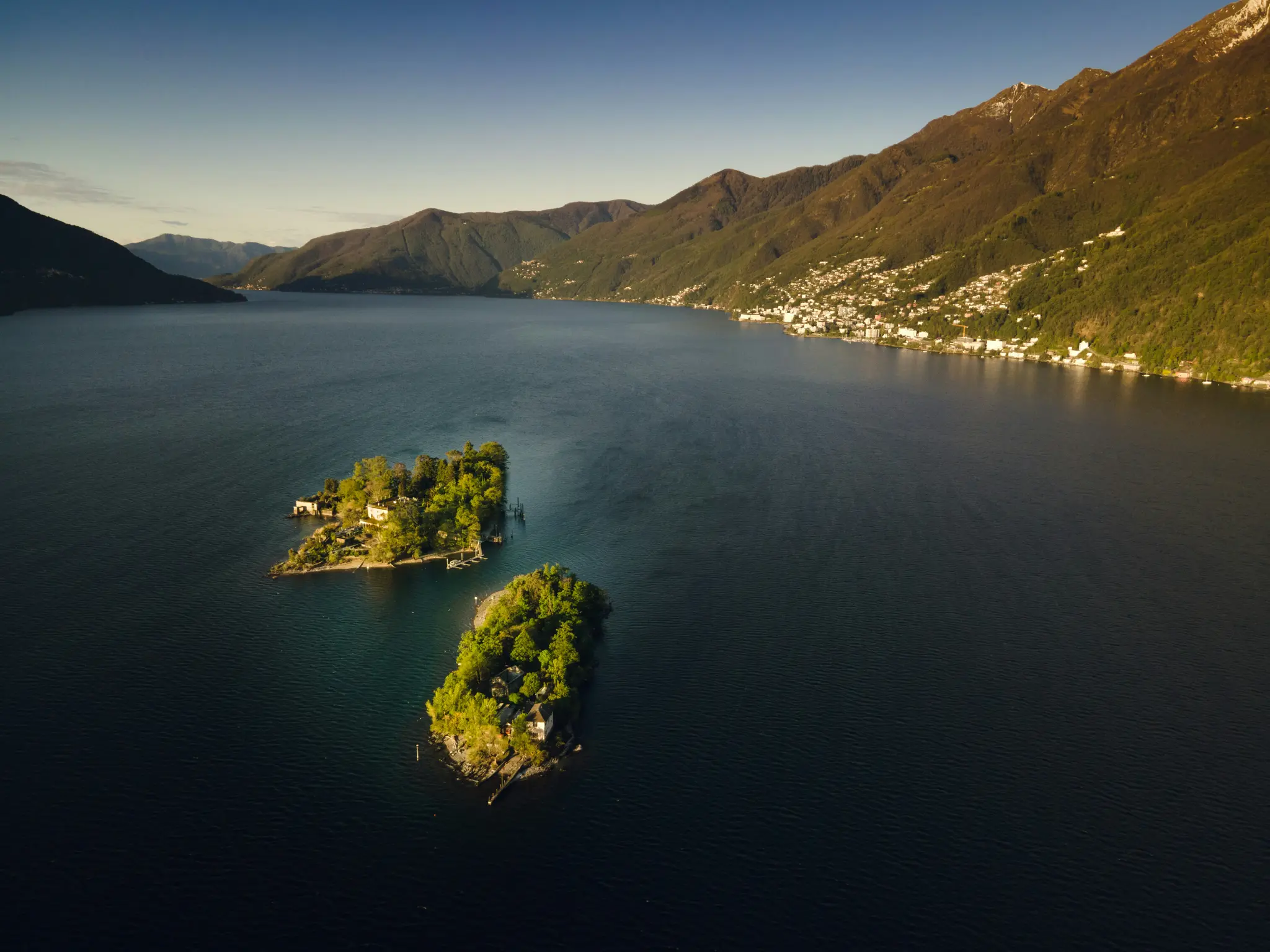 En flygfoto av två små, gröna öar med hus i en mörkblå sjö. Berg reser sig på båda sidor, med en stad på…