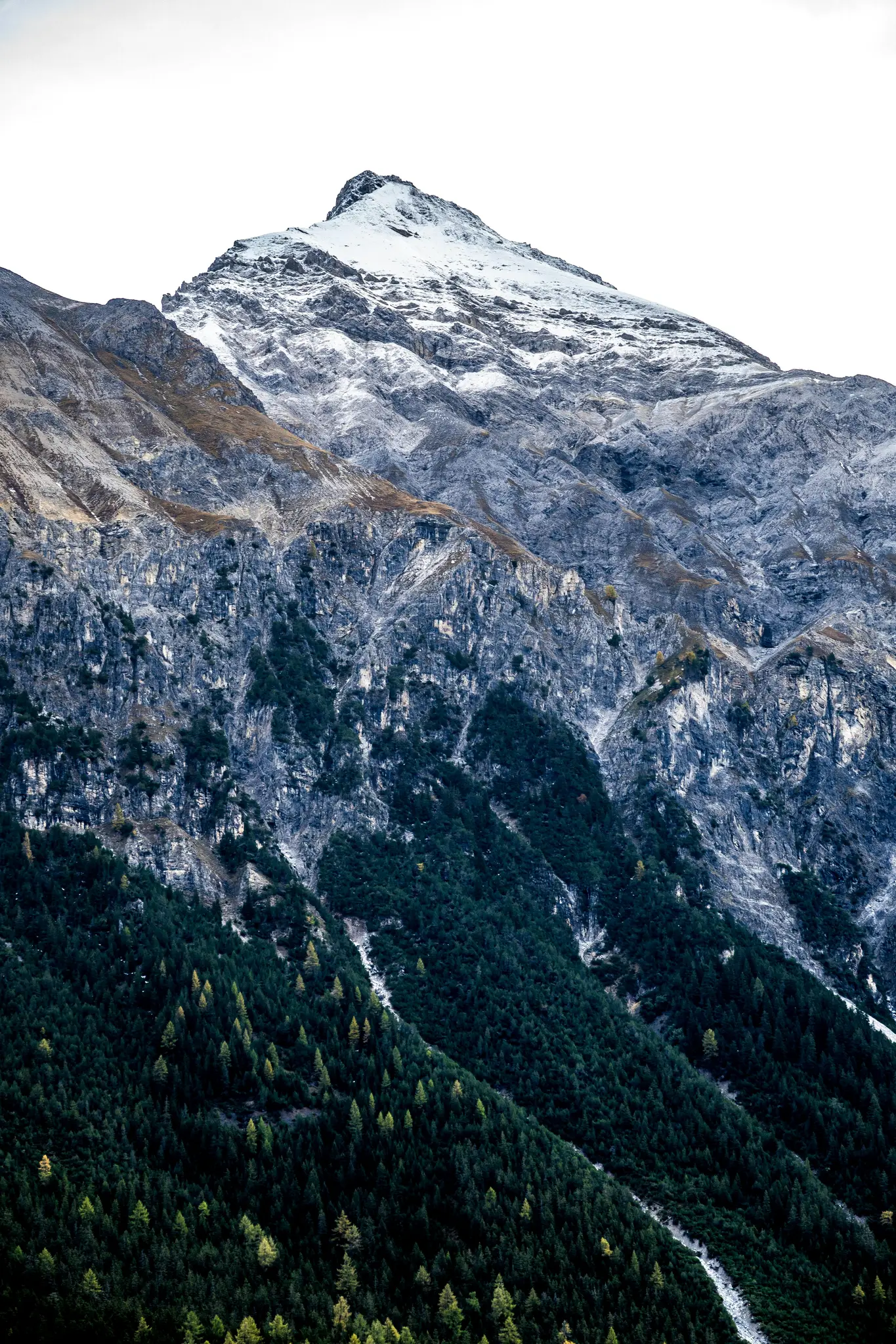 En hög, snöklädd bergstopp dominerar den övre delen av bilden, med steniga grå sluttningar under den. Den nedre halvan…