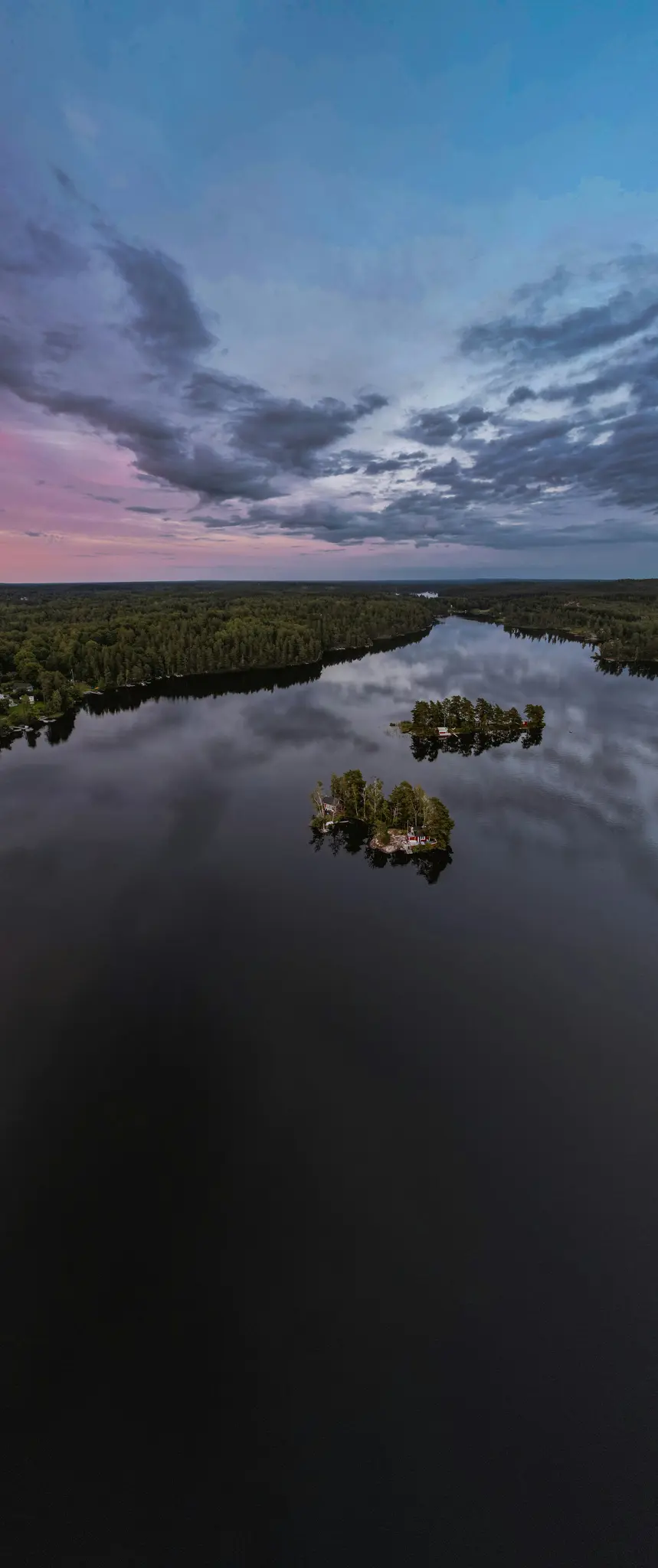 En flygbild av en mörk sjö som reflekterar en molnig himmel, med flera små, trädklädda öar och en tät skog längs…