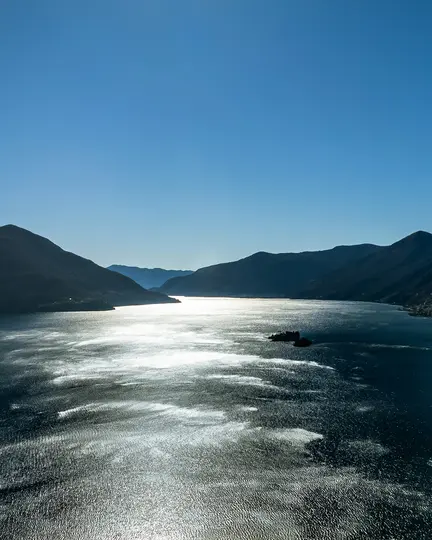 Una vista aérea muestra un vasto lago con luz solar reluciente reflejada en su superficie, flanqueado por montañas oscuras