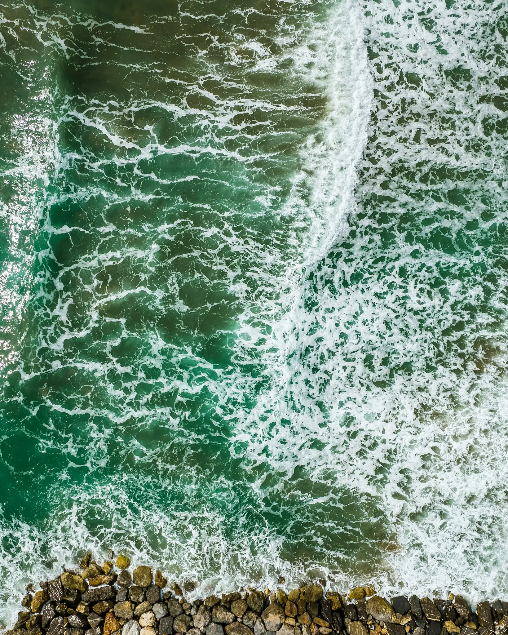Una vista aérea muestra vibrantes olas de océano verdes y azules con espuma blanca rompiendo contra una costa oscura y