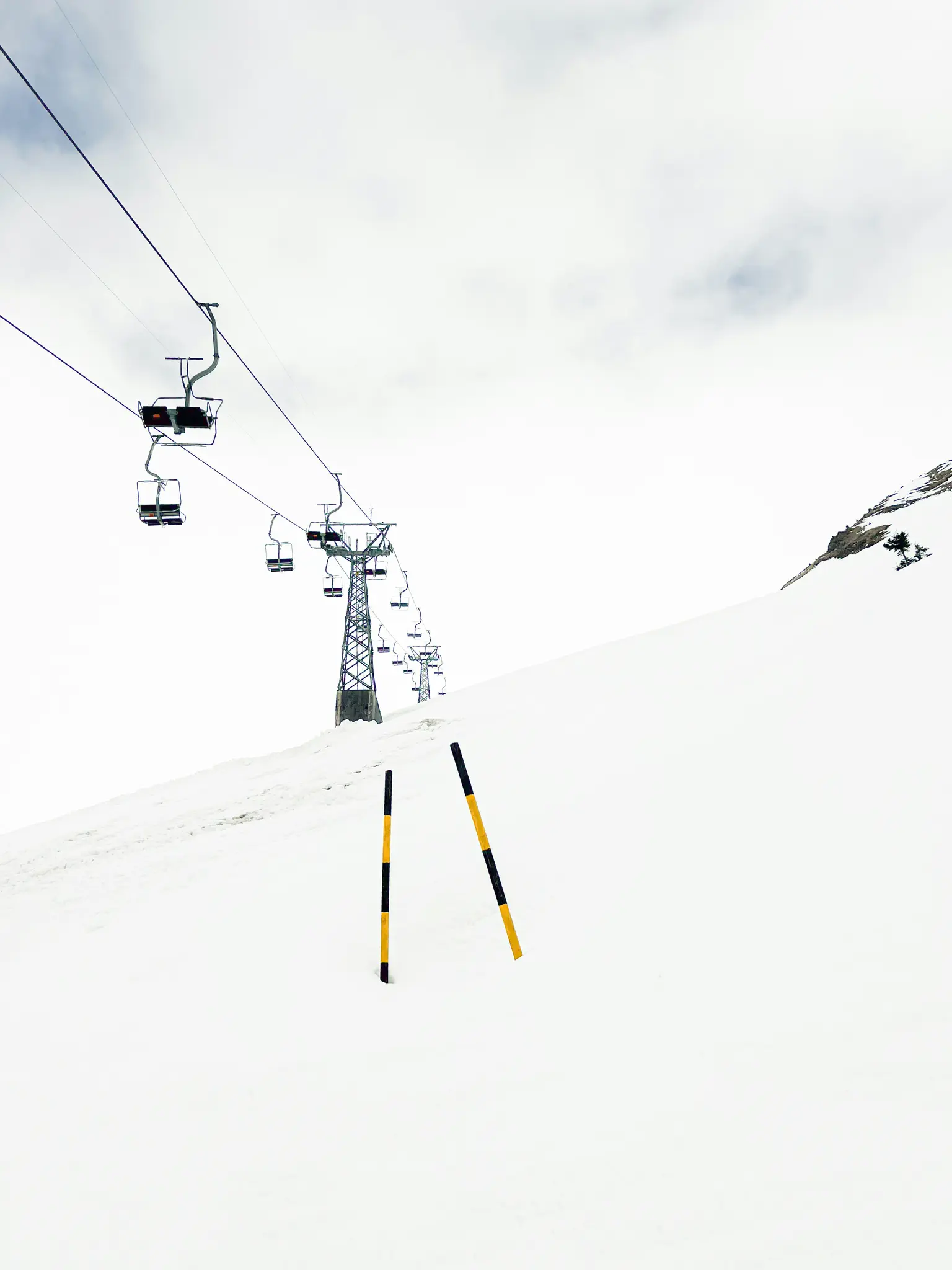 An empty ski lift with multiple chairs ascends a vast, snow-covered mountain slope under a cloudy, white sky. Two yellow…