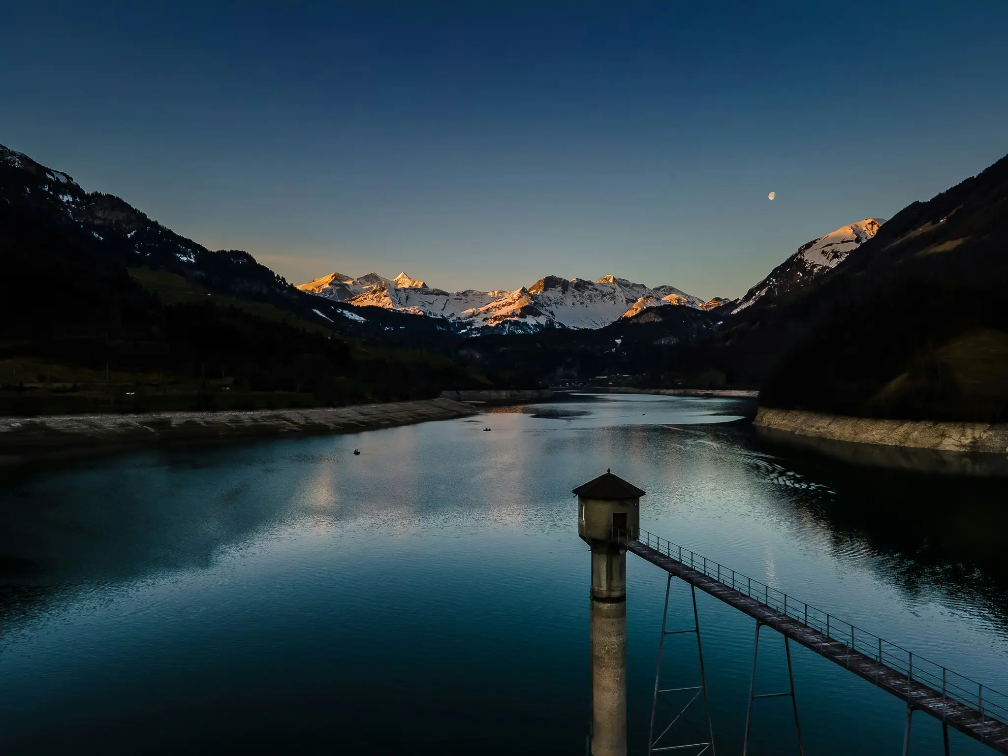 A wide landscape photo of a calm mountain lake at twilight, with a small tower on a pier extending into the water.…
