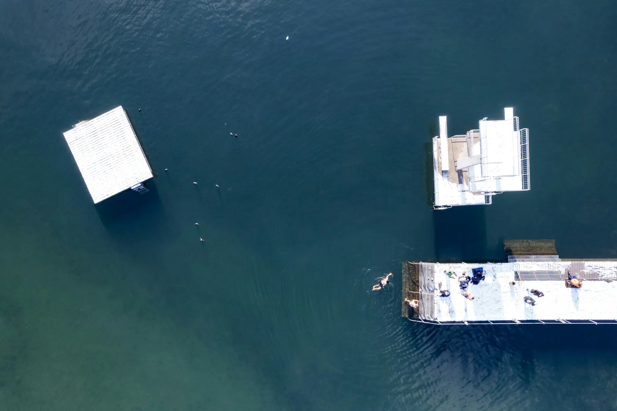 Una toma aérea muestra un largo muelle blanco y dos plataformas flotantes blancas sobre agua azul oscuro. Hay personas en el