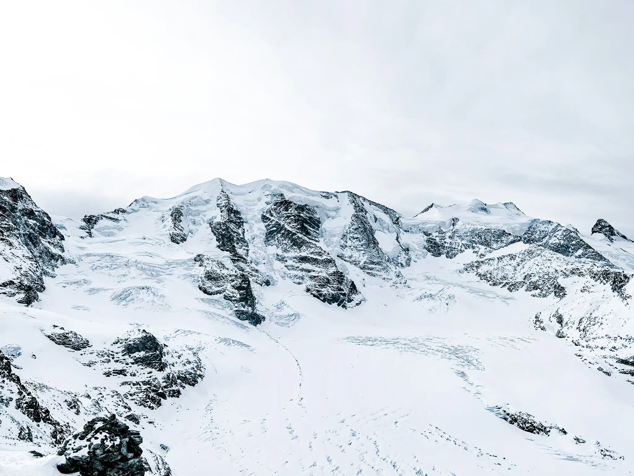 En vidvinkelbild av en majestätisk, snötäckt bergskedja under en ljus, molnig himmel. Glaciärer är synliga.