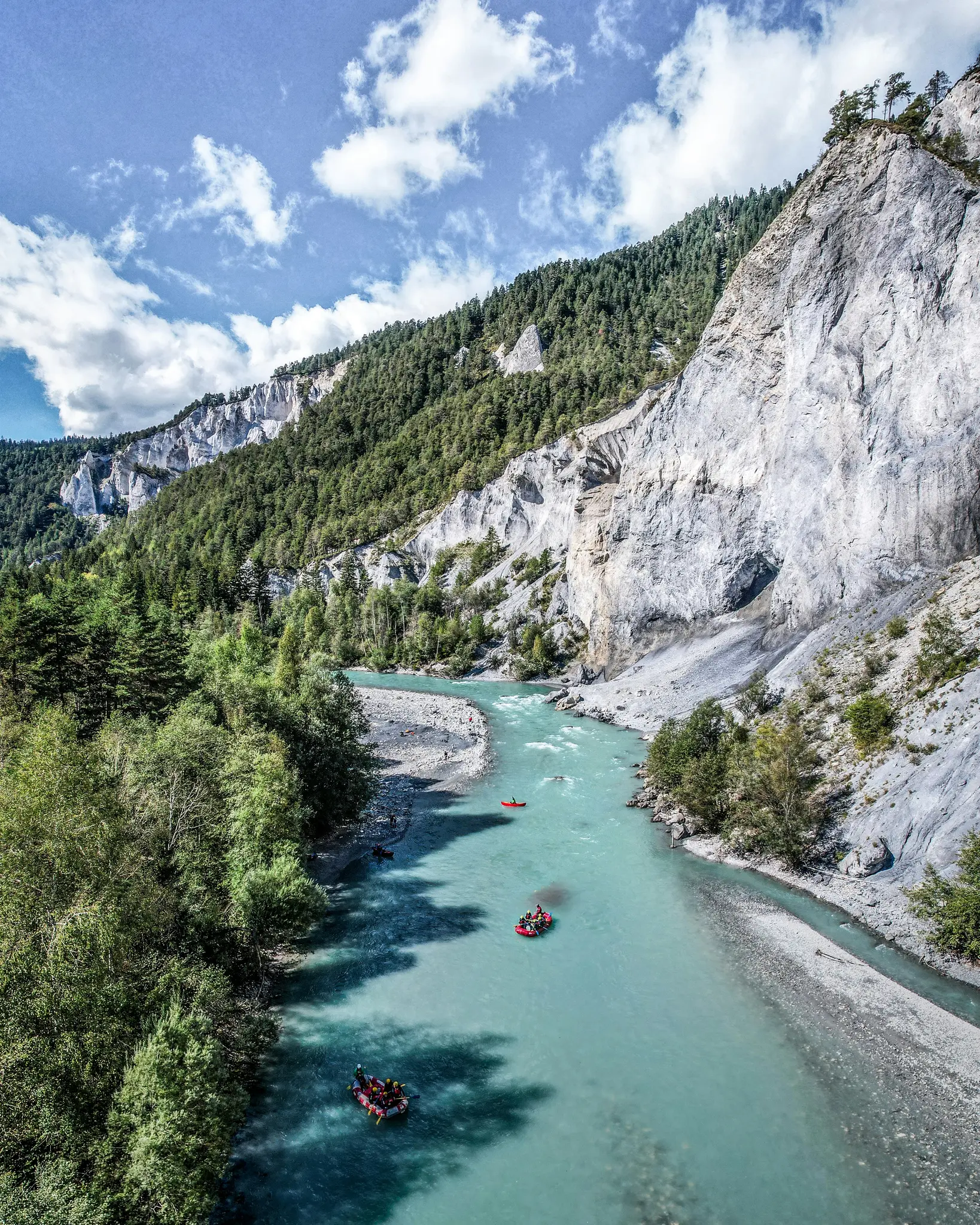 Aerial view of a turquoise river flowing through a deep valley with lush green forests and towering grey cliffs. Several…