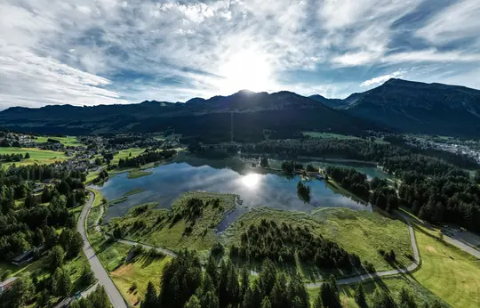 Ett brett landskapsfoto visar en sjö som reflekterar solen och himlen, omgiven av gröna skogar, fält och en stad.